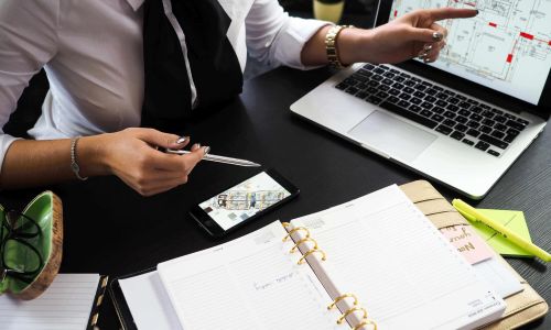 A high-angle shot of a person in a white shirt and black tie working at a desk. One hand points to an architectural floor plan on a laptop screen, while the other holds a pen over a smartphone displaying a similar plan. An open daily planner, glasses, and a highlighter are also visible on the black desk surface.